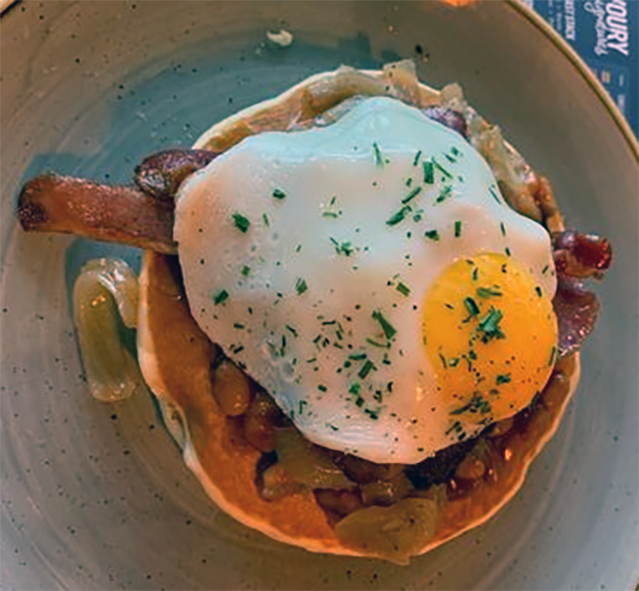 pile of scotch pancakes with bacon, tomatoes topped with a fried egg