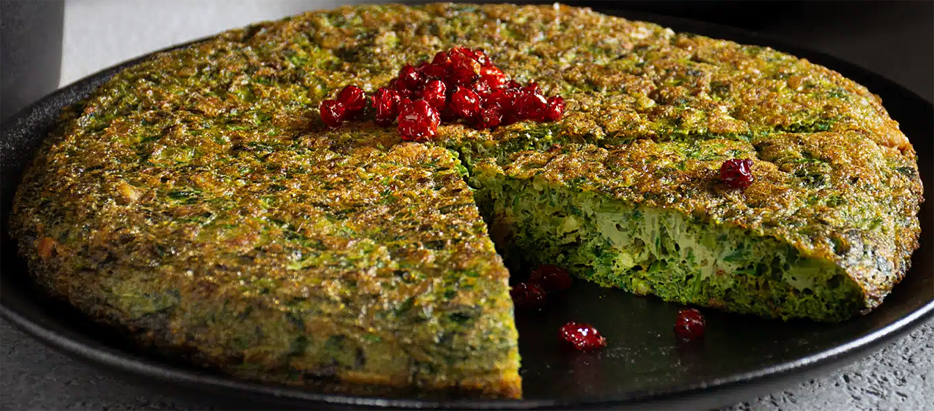 kuku sabzi, Persian herb frittata served on a plate garnished with dried barberries and with a wedge removed to reveal the inside