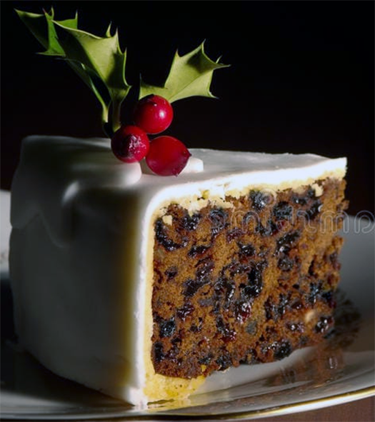 slice of rich Christmas cake with marzipan and icing garnished with a sprig of holly on a plate