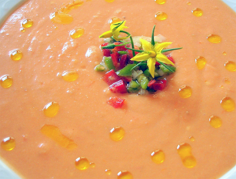cold tomato soup in a bowl garnished with olive oil, finely chopped vegetables and flowers