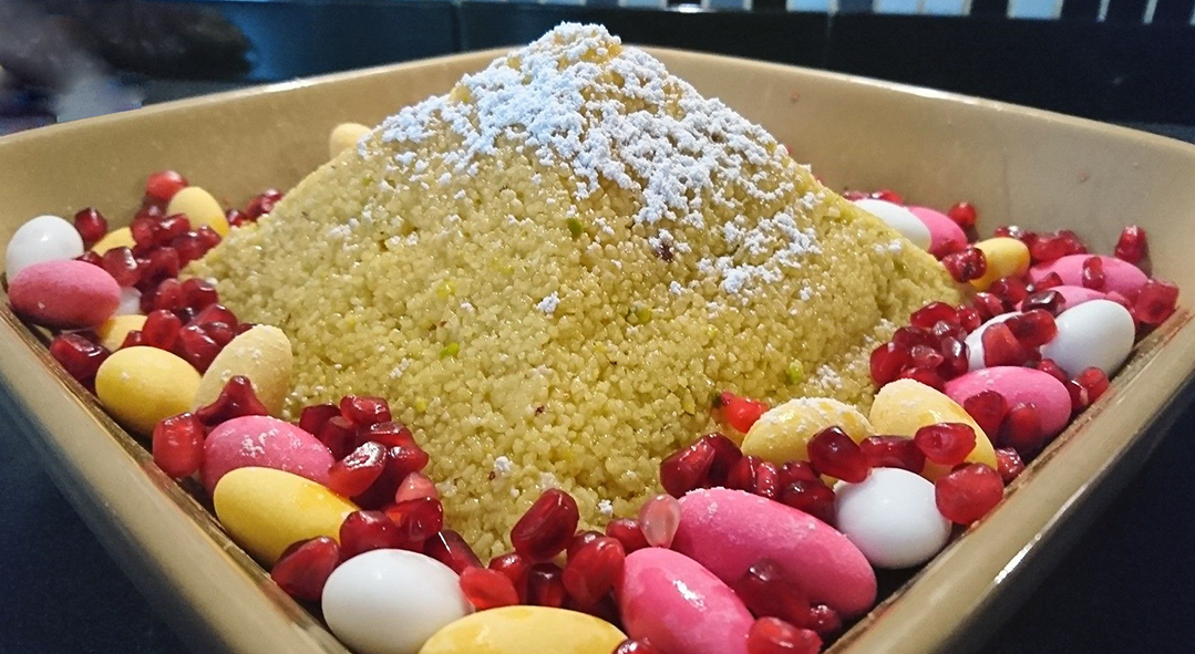pyramid of sweet couscous topped with powdered sugar served surrounded by candied almonds and pomegranate seeds