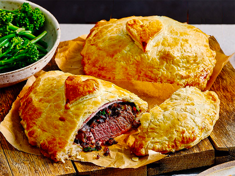 two individual beef wellingtons on a chopping board next to a bowl of broccoli, with one sliced to reveal the beef inside