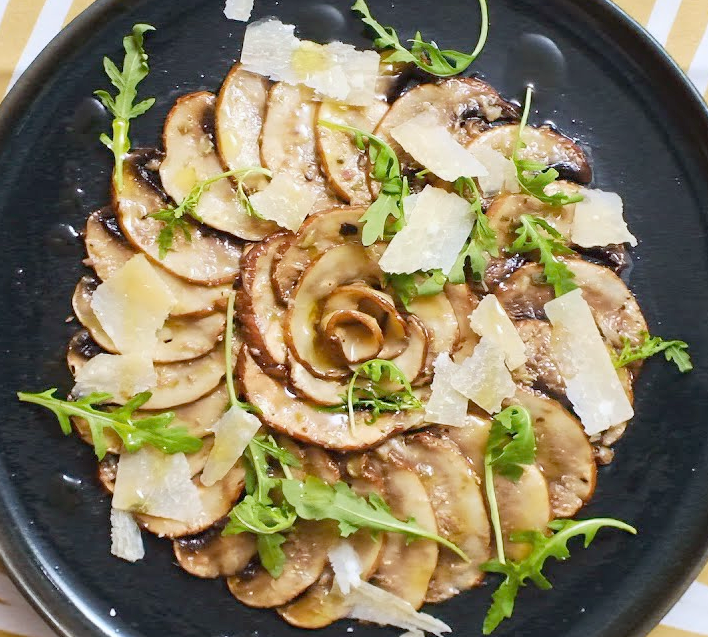 carpaccio of thinly-sliced st georges mushrooms arranged decoratively on a plate and garnished with olive oil, rocket leaves and parmesan shavings
