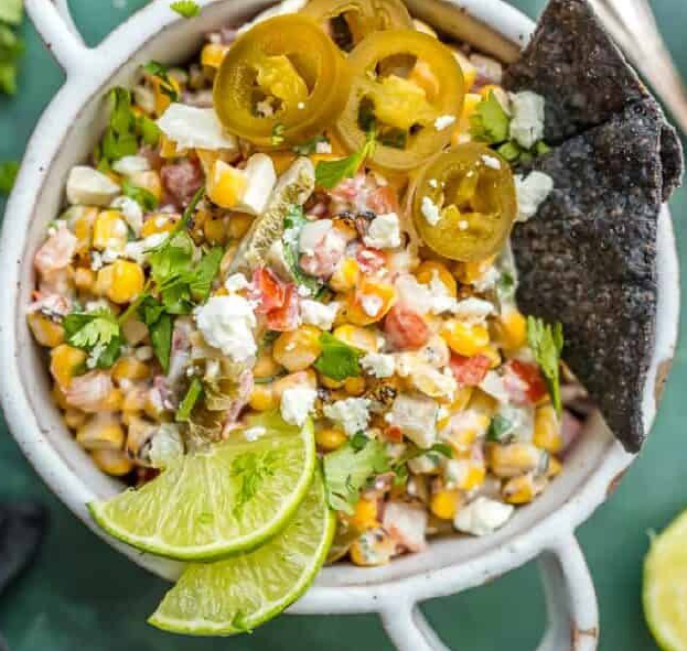 street corn salsa in a bowl garnished with lime wedges, tortilla chips and pickled chillies