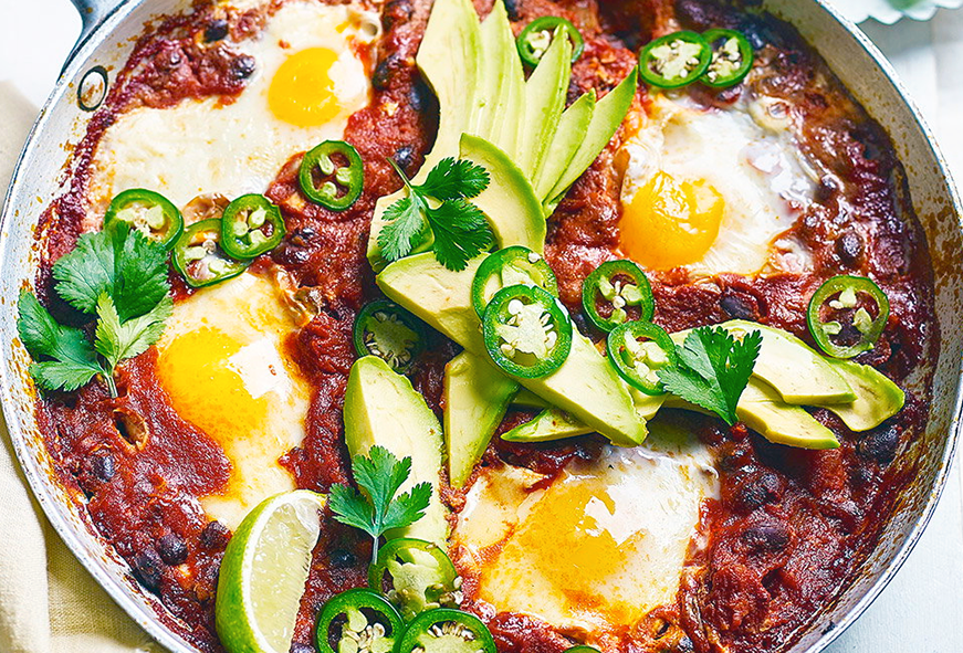 black beans huzvos rancheros with four eggs in a frying pan served topped with sliced avocado and sliced chillies