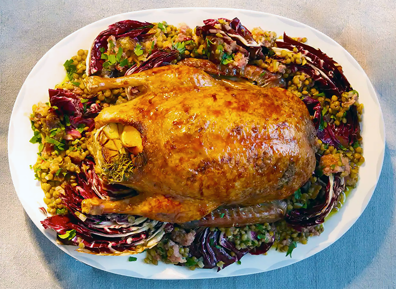 Roast duck stuffed with oranges served on a plate surrounded by a mix of lentils, cotechino sausage, dates, radicchio and parsley
