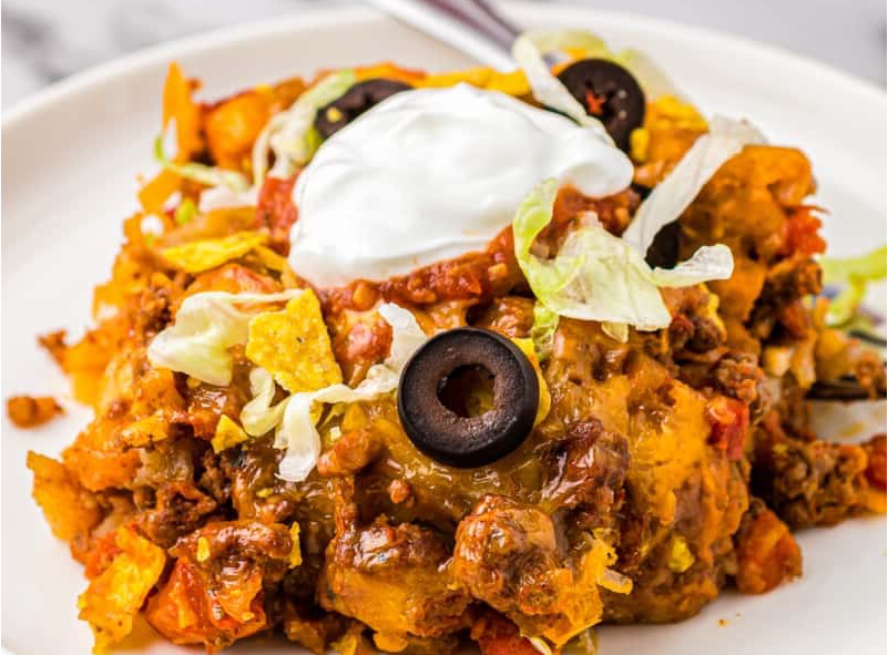 spoonful of taco casserole, a blend of minced beef, tater tots, refried beans, and lots of cheese served on a plate with a garnish of sour cream