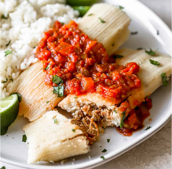 two tamales served on a plate with rice and a garnish of tomato salsa, one split open to reveal the filling